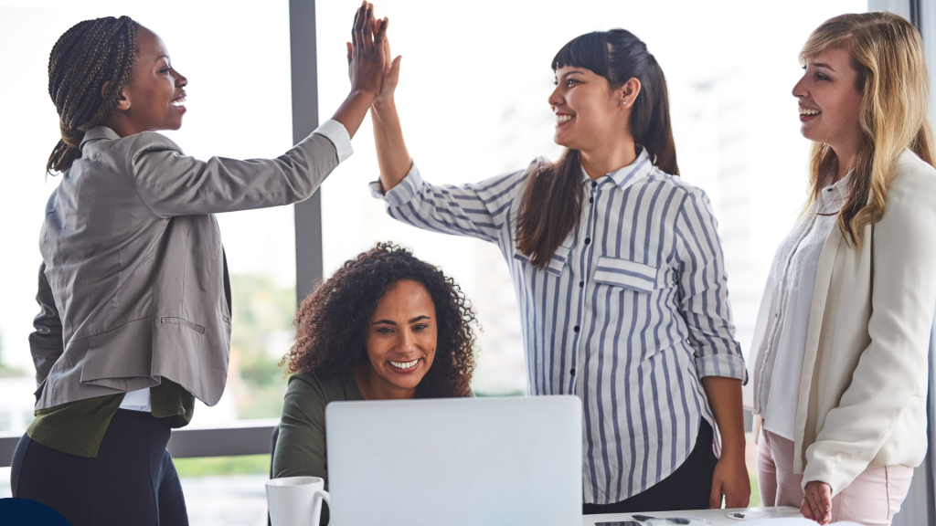Four women in a modern office space celebrate together. Two of them are giving a high-five, while another smiles at a laptop. The mood is positive and collaborative. Quatre femmes dans un bureau moderne célèbrent ensemble. Deux d’entre elles se donnent un high-five, tandis qu’une autre sourit devant un ordinateur portable. L’ambiance est positive et collaborative.