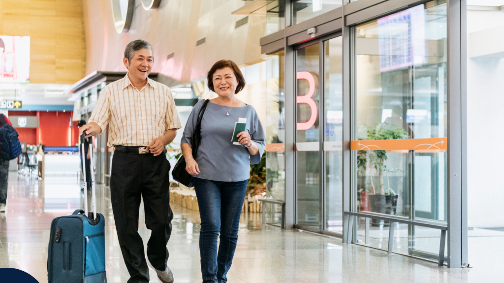 An older couple walks through an airport terminal smiling. One person pulls a suitcase while the other holds travel documents. They appear relaxed and ready for their journey. Un couple âgé marche en souriant dans un terminal d’aéroport. L’un tire une valise tandis que l’autre tient des documents de voyage. Ils semblent détendus et prêts pour leur voyage.