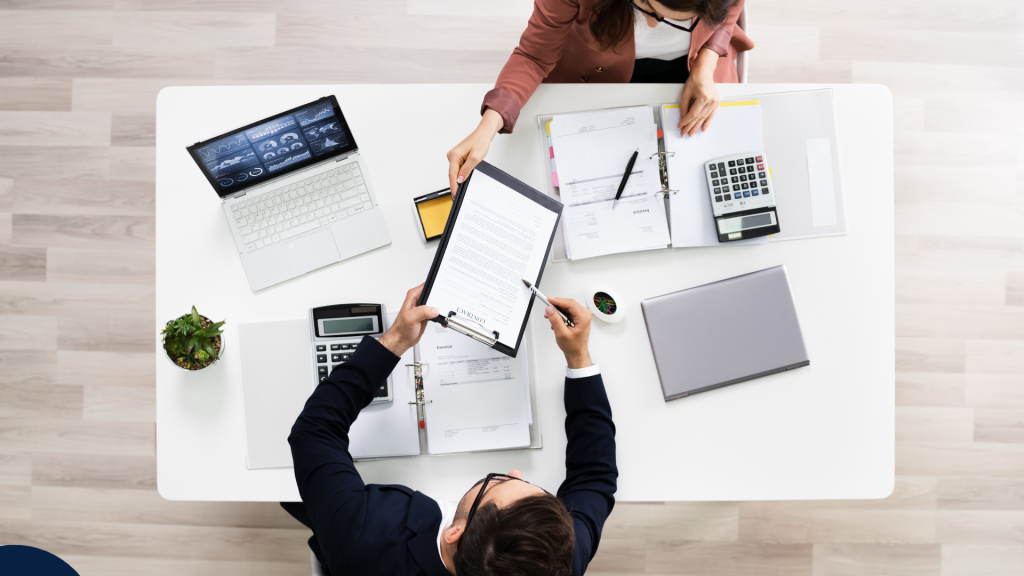 Two people sit across from each other at a white desk, exchanging a clipboard. The table has laptops, documents, a calculator, and coffee cups, suggesting a meeting or business discussion. Deux personnes sont assises face à face à un bureau blanc et s’échangent une planchette à pince. Sur la table se trouvent des ordinateurs portables, des documents, une calculatrice et des tasses de café, suggérant une réunion ou une discussion d’affaires.