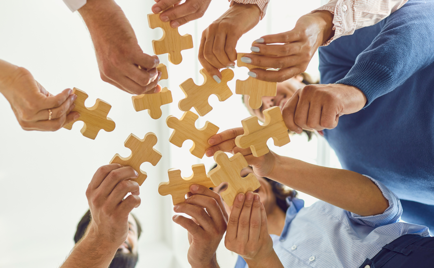 Multiple people raise their hands in a circle, each holding a large wooden puzzle piece. The group appears diverse, symbolizing teamwork, unity, and collaboration. Plusieurs personnes lèvent les mains en cercle, chacune tenant une grande pièce de casse-tête en bois. Le groupe est diversifié, symbolisant le travail d’équipe, l’unité et la collaboration.