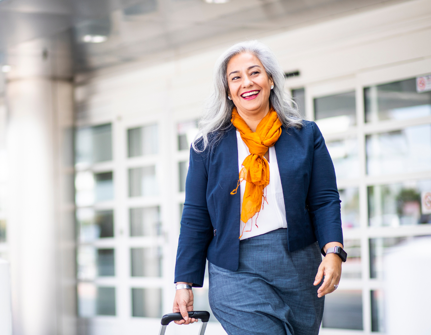 A smiling woman with long gray hair walks confidently while pulling a suitcase. She’s dressed in business-casual clothing and wears a bright orange scarf. The setting is outside an airport. Une femme souriante aux longs cheveux gris marche avec assurance en tirant une valise. Elle porte des vêtements de style professionnel-décontracté et une écharpe orange vif. La scène se déroule à l’extérieur d’un aéroport.