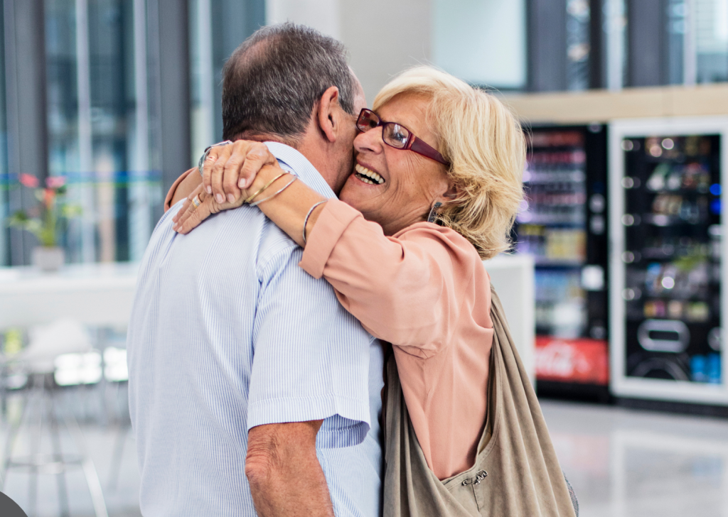 An older woman with glasses warmly hugs an older man in an airport space. She’s smiling joyfully, and they appear happy to see each other, possibly reunited after time apart. Une femme âgée portant des lunettes serre chaleureusement un homme âgé dans ses bras, dans un aéroport. Elle sourit avec joie. Ils semblent heureux de se retrouver, peut-être après une longue séparation.