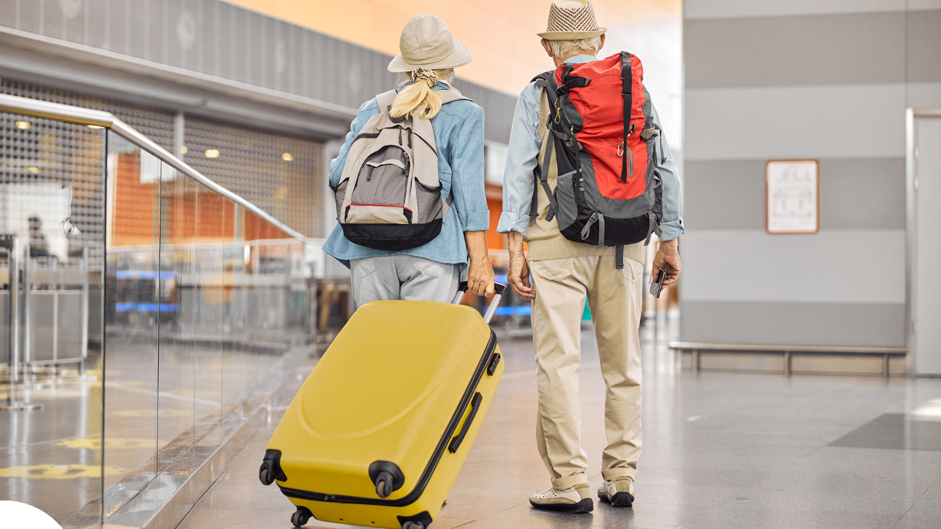 An older couple walks side by side through a modern airport terminal. Both are wearing backpacks and sun hats. The person on the left is pulling a large, bright yellow suitcase. Their casual clothing and relaxed pace suggest they are seasoned travelers on vacation. Un couple âgé marche côte à côte dans un terminal d’aéroport moderne. Les deux portent des sacs à dos et des chapeaux de soleil. La personne à gauche tire une grande valise jaune vif. Leurs vêtements décontractés et leur allure tranquille suggèrent qu’ils sont des voyageurs expérimentés en vacances.