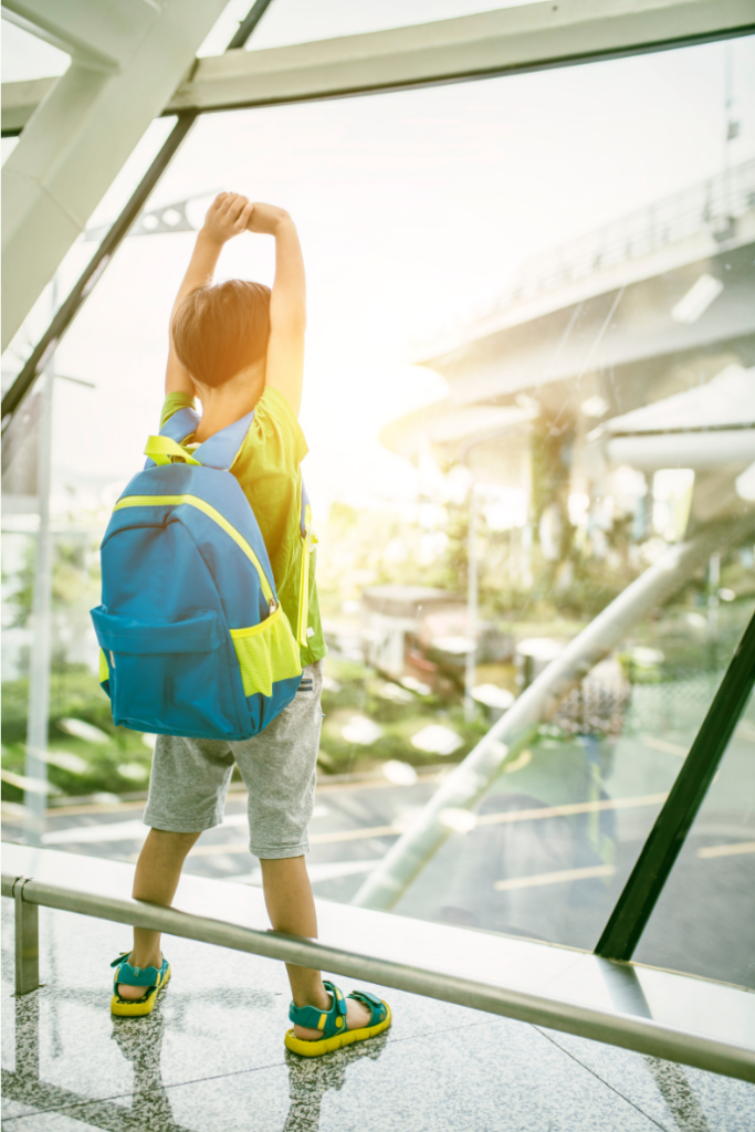 A young child with a bright blue and green backpack stands by a large window at an airport, stretching their arms up while looking outside at a sunny urban landscape. Un jeune enfant portant un sac à dos bleu et vert vif se tient près d’une grande fenêtre dans un aéroport. Il lève les bras en regardant à l’extérieur vers un paysage urbain ensoleillé.