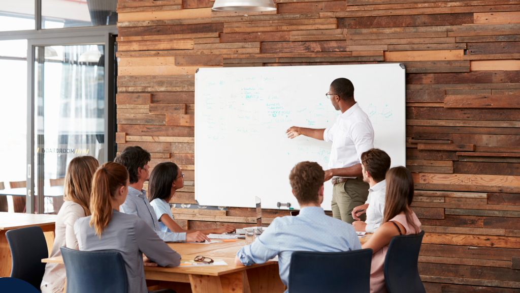 A man stands at a whiteboard presenting to a group of seated colleagues in a modern meeting room with wooden walls. The group appears focused, suggesting a team discussion or training session. Un homme se tient devant un tableau blanc et fait une présentation à un groupe de collègues assis dans une salle de réunion moderne aux murs en bois. Le groupe semble concentré, suggérant une discussion d’équipe ou une séance de formation.