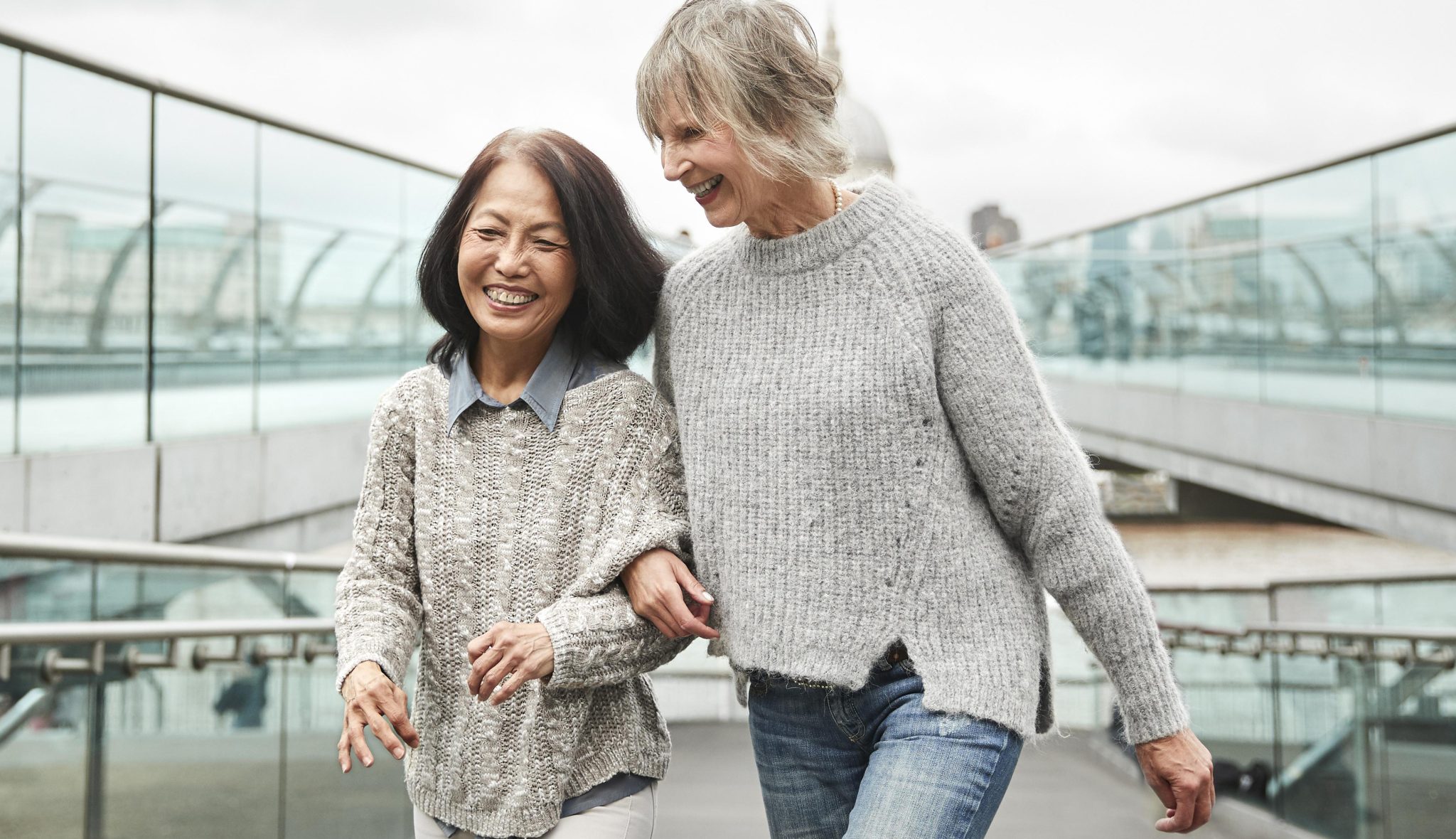 Two older women are walking arm-in-arm on a modern pedestrian bridge, smiling and laughing. They look relaxed and happy, wearing cozy sweaters on a cloudy day. Deux femmes âgées marchent bras dessus bras dessous sur une passerelle piétonne moderne. Elles sourient et rient, portant des chandails confortables par une journée nuageuse. Elles ont l’air détendues et heureuses.