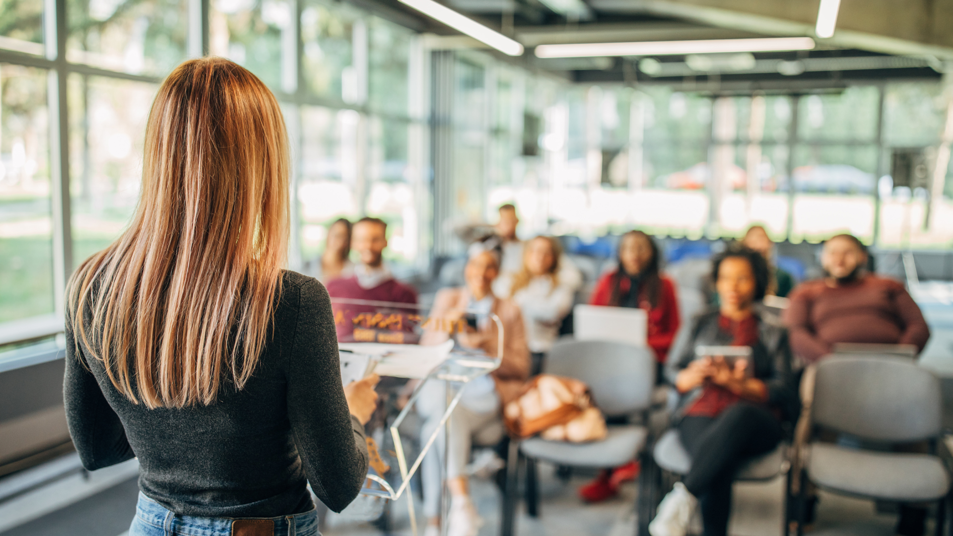 A woman with long hair stands at a podium speaking to a seated group in a bright, modern room with large windows. The audience looks engaged, suggesting a lecture, workshop, or presentation setting. Une femme aux longs cheveux se tient à un podium et s’adresse à un groupe assis dans une salle moderne et lumineuse avec de grandes fenêtres. Le public semble attentif, suggérant un contexte de conférence, d’atelier ou de présentation.