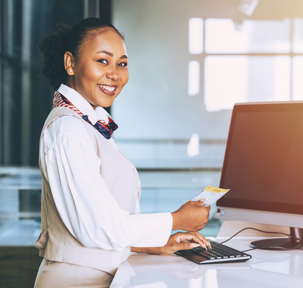 A smiling airport staff member stands at a check-in counter holding a boarding pass and typing on a keyboard. She wears a white uniform with a red scarf and looks welcoming and professional. Une employée d’aéroport souriante se tient au comptoir d’enregistrement, tenant une carte d’embarquement et tapant sur un clavier. Elle porte un uniforme blanc avec un foulard rouge et a une apparence accueillante et professionnelle.